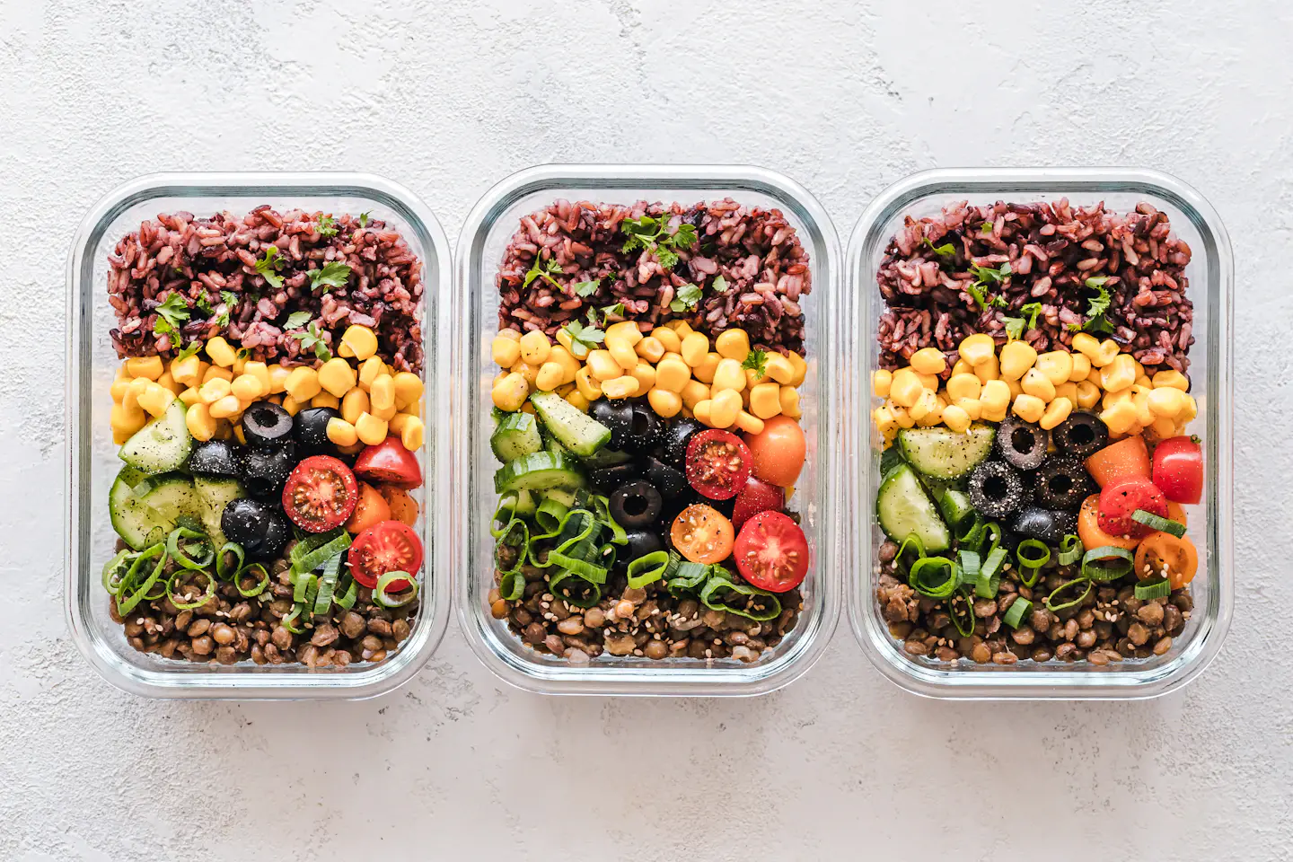 Three glass meal-prep containers with rice, lentils, corn, olives, and chopped vegetables.